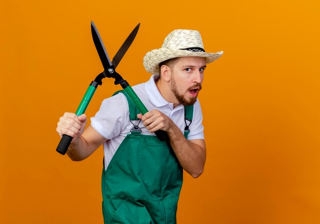 Beeindruckter junger hübscher slawischer Gärtner in Uniform und Hut, die Scheren halten, die isoliert schauen