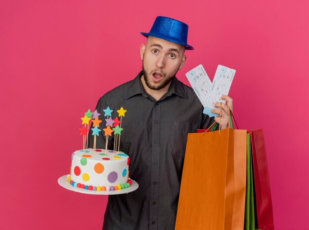 Beeindruckter junger hübscher slawischer Party-Typ, der Partyhut hält, der Geburtstagskuchen-Flugtickets und Papiertüten betrachtet, die Kamera lokal auf purpurrotem Hintergrund betrachten