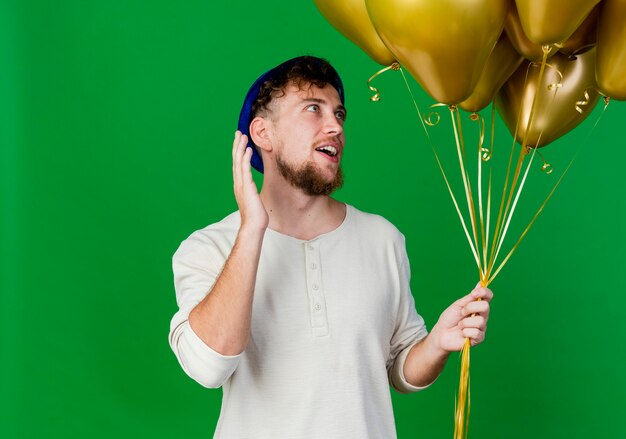Beeindruckter junger hübscher slawischer Party-Typ, der Partyhut hält und Ballons hält, die Hand in der Luft lokalisiert auf grünem Hintergrund mit Kopienraum halten