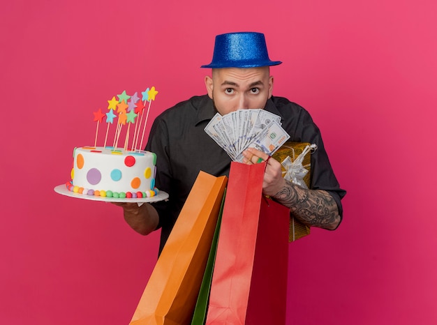 Beeindruckter junger hübscher slawischer Party-Typ, der Partyhut trägt, der Geldpapiertüten-Geschenkpackung und Geburtstagstorte hält und Kamera betrachtet, die auf purpurrotem Hintergrund mit Kopienraum lokalisiert wird