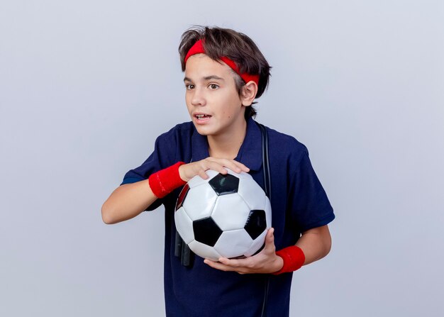 Beeindruckter junger hübscher sportlicher Junge, der Stirnband und Armbänder mit Zahnspangen und Springseil um Hals hält, der Fußball hält, der gerade lokalisiert auf weißem Hintergrund mit Kopienraum schaut