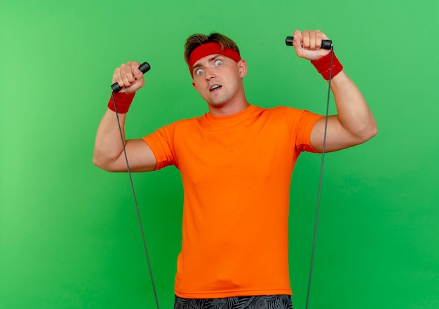 Beeindruckter junger hübscher sportlicher Mann, der Stirnband und Armbänder trägt, die Springseil hochziehen, lokalisiert auf Grün