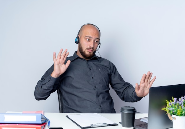 Beeindruckter junger kahlköpfiger Callcenter-Mann, der ein Headset trägt, das am Schreibtisch mit Arbeitswerkzeugen sitzt, die Laptop betrachten und leere Hände zeigen, die Stoppgeste am Laptop lokalisiert auf Weiß tun