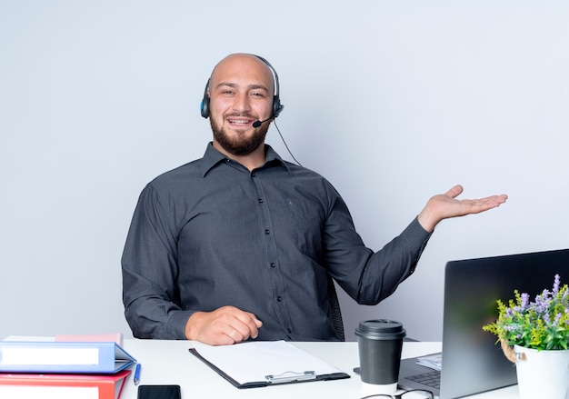 Beeindruckter junger kahlköpfiger Callcenter-Mann, der Headset trägt, das am Schreibtisch mit Arbeitswerkzeugen sitzt, die leere Hand lokalisiert auf Weiß zeigen