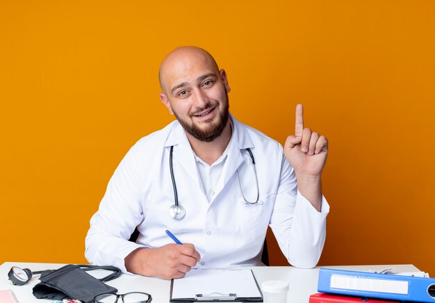 Beeindruckter junger kahlköpfiger männlicher Arzt, der medizinische Robe und Stethoskop trägt und am Schreibtisch sitzt sitting