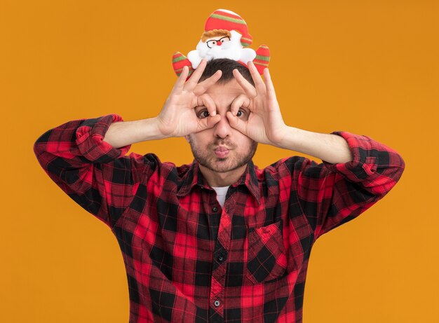 Beeindruckter junger kaukasischer Mann, der das Weihnachtsmann-Stirnband trägt, das Kamera betrachtet, die Blickgeste unter Verwendung der Hände als Fernglas mit gespitzten Lippen lokalisiert auf orangeem Hintergrund tut