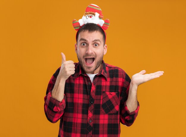 Beeindruckter junger kaukasischer Mann, der das Weihnachtsmann-Stirnband trägt, das Kamera betrachtet, die Daumen oben und leere Hand lokalisiert auf orange Hintergrund zeigt