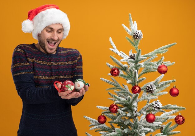 Beeindruckter junger kaukasischer Mann, der Weihnachtshut trägt, der nahe dekoriertem Weihnachtsbaum hält und Weihnachtskugeln lokalisiert auf orangefarbenem Hintergrund hält