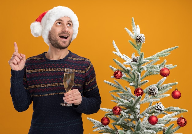 Beeindruckter junger kaukasischer Mann, der Weihnachtshut trägt, der nahe verziertem Weihnachtsbaum steht, der Glas Champagner hält und lokalisiert auf orange Wand hält