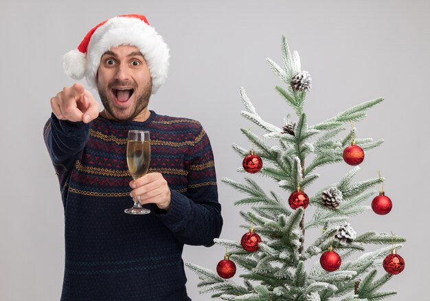 Beeindruckter junger kaukasischer Mann, der Weihnachtshut trägt, der nahe Weihnachtsbaum hält, der Glas Champagner hält und lokalisiert auf weißer Wand zeigt