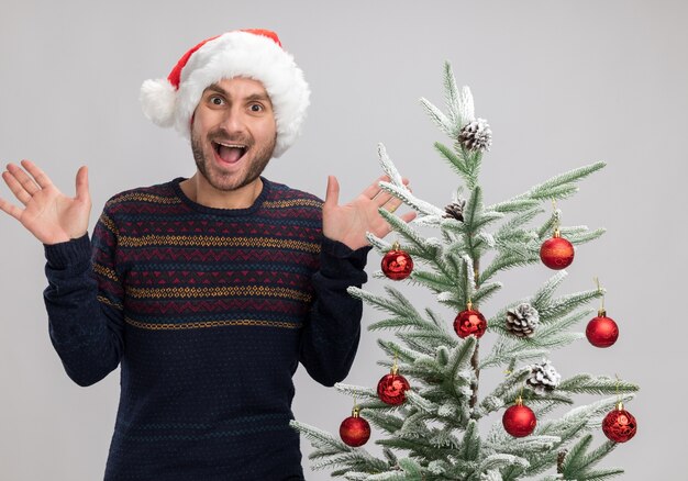 Beeindruckter junger kaukasischer Mann, der Weihnachtshut trägt, der nahe Weihnachtsbaum steht und Kamera betrachtet, die leere Hände lokalisiert auf weißem Hintergrund zeigt