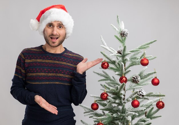 Beeindruckter junger kaukasischer Mann, der Weihnachtshut trägt, der nahe Weihnachtsbaum steht und Kamera betrachtet, die mit Händen auf Baum lokalisiert auf weißem Hintergrund zeigt