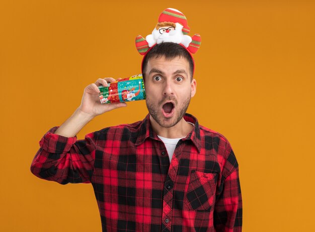 Beeindruckter junger kaukasischer Mann, der Weihnachtsmann-Stirnband hält, das Plastikweihnachtsbecher neben Ohr hält, das Kamera lokalisiert auf orangefarbenem Hintergrund betrachtet