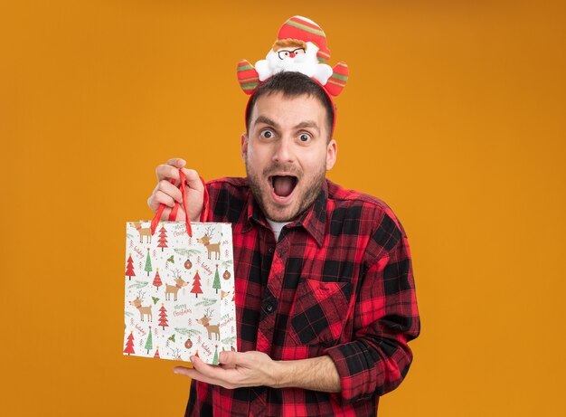 Beeindruckter junger kaukasischer Mann, der Weihnachtsmann-Stirnband hält, das Weihnachtsgeschenkbeutel lokalisiert auf orange Wand mit Kopienraum hält