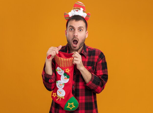 Beeindruckter junger kaukasischer Mann, der Weihnachtsmann-Stirnband hält, das Weihnachtsstrumpf hält, der es lokalisiert auf orange Wand mit Kopienraum öffnet