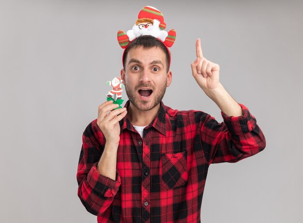 Beeindruckter junger kaukasischer Mann, der Weihnachtsmann-Stirnband hält Schneemann-Weihnachtsspielzeug, das Kamera zeigt, die oben auf weißem Hintergrund zeigt