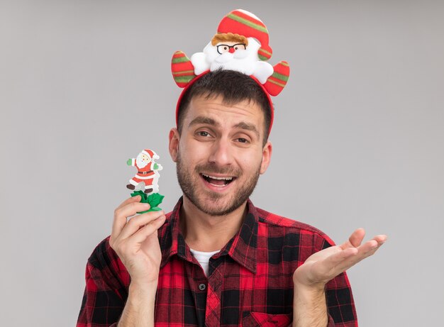Beeindruckter junger kaukasischer Mann, der Weihnachtsmann-Stirnband hält Schneemann-Weihnachtsspielzeug zeigt, das leere Hand lokalisiert auf weißer Wand zeigt