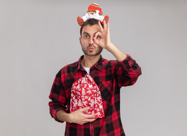 Beeindruckter junger kaukasischer Mann, der Weihnachtsstirnband hält, das Weihnachtssack tut, Blick schauen Geste mit gespitzten Lippen lokalisiert auf weißer Wand mit Kopienraum