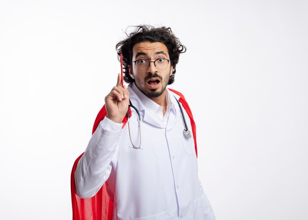 Beeindruckter junger kaukasischer Superheldenmann in optischer Brille in Arztuniform mit rotem Mantel und mit Stethoskop um den Hals hält Bleistift neck