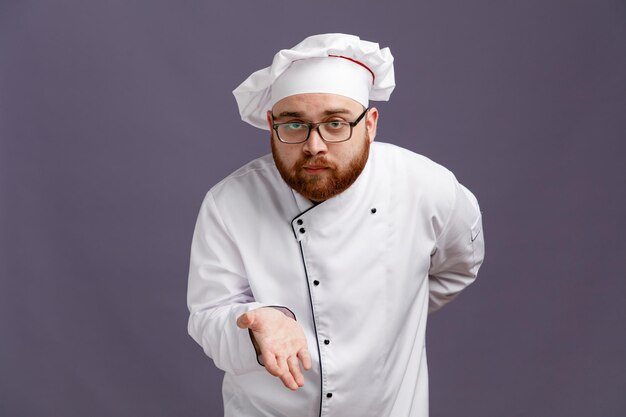 Beeindruckter junger Koch mit Brille, Uniform und Mütze, der in die Kamera blickt und eine leere Hand zeigt, während er eine andere Hand hinter seinem Rücken isoliert auf violettem Hintergrund hält