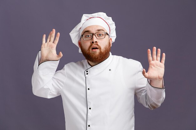 Beeindruckter junger Koch mit einheitlicher Brille und Mütze, der in die Kamera blickt und leere Hände isoliert auf violettem Hintergrund zeigt