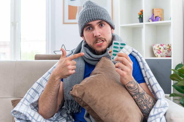 Beeindruckter junger kranker Mann mit Schal und Wintermütze, der in eine Decke gehüllt ist, sitzt auf dem Sofa im Wohnzimmer und hält Kissen und Pillenpackungen mit Blick auf die Kamera, die auf Pillen zeigt