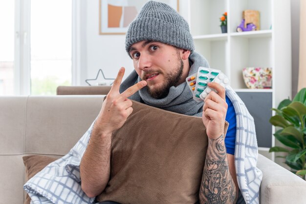 Beeindruckter junger kranker Mann mit Schal und Wintermütze, der in eine Decke gehüllt ist und auf dem Sofa im Wohnzimmer sitzt und ein Kissen hält, das in die Kamera schaut, die Pillenpackungen zeigt und zwei mit der Hand zeigt