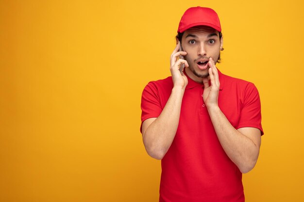 Beeindruckter junger Lieferbote in Uniform und Mütze, der in die Kamera blickt und die Hand in der Nähe des Mundes hält, der am Telefon isoliert auf gelbem Hintergrund mit Kopierraum spricht