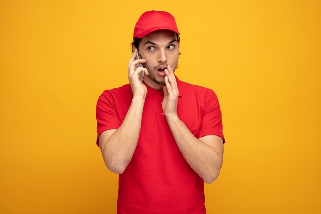 Beeindruckter junger Lieferbote in Uniform und Mütze, der zur Seite schaut und die Hand in der Nähe des Mundes hält, der am Telefon isoliert auf gelbem Hintergrund flüstert