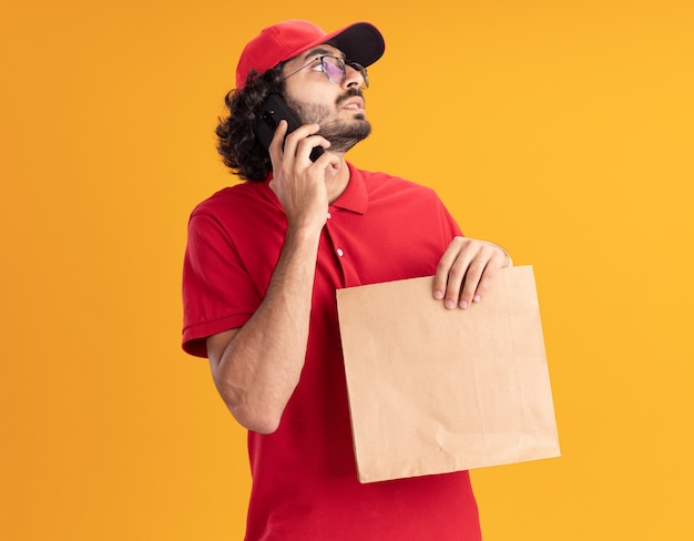 Beeindruckter junger liefermann in roter uniform und mütze mit brille, die ein papierpaket hält, das am telefon spricht und auf die seite isoliert auf der orangefarbenen wand schaut