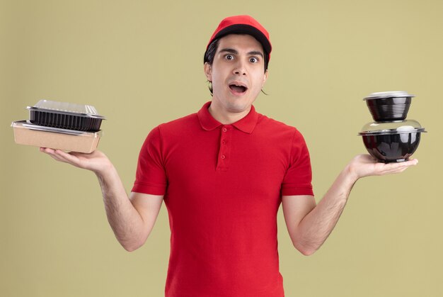 Beeindruckter junger Liefermann in roter Uniform und Mütze mit Lebensmittelbehältern und Papierverpackungen mit Blick auf die Vorderseite isoliert auf olivgrüner Wand