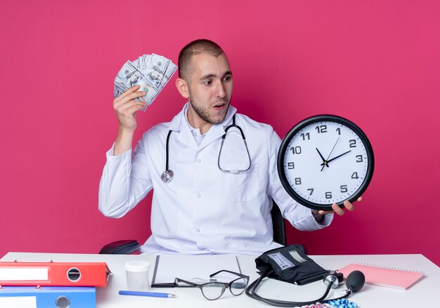 Beeindruckter junger männlicher Arzt, der medizinische Robe und Stethoskop trägt, die am Schreibtisch mit Arbeitswerkzeugen sitzen, die Uhr und Geld halten, die auf Rosa lokalisiert betrachten