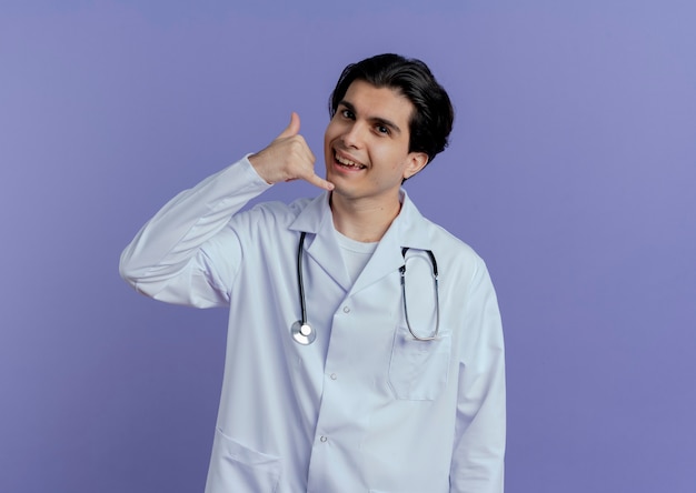 Beeindruckter junger männlicher Arzt, der medizinisches Gewand und Stethoskop trägt, die Rufgeste lokalisiert auf lila Wand mit Kopienraum tun