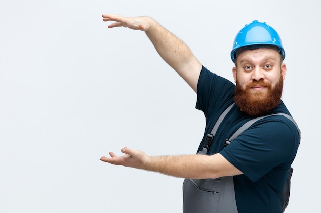 Kostenloses Foto beeindruckter junger männlicher bauarbeiter mit schutzhelm und uniform, der in der profilansicht steht und in die kamera blickt und eine große geste zeigt, die auf weißem hintergrund mit kopierraum isoliert ist