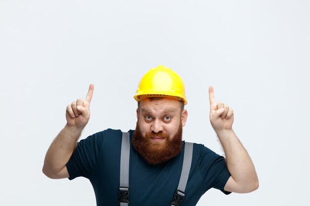 Beeindruckter junger männlicher Bauarbeiter mit Schutzhelm und Uniform, der in die Kamera blickt und mit dem Finger nach oben zeigt, isoliert auf weißem Hintergrund mit Kopierraum