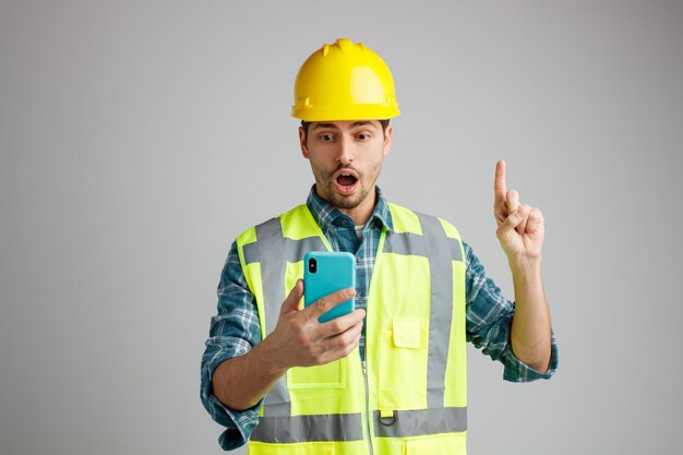Beeindruckter junger männlicher Ingenieur mit Schutzhelm und Uniform in der Hand und Blick auf das Handy, das isoliert auf weißem Hintergrund nach oben zeigt