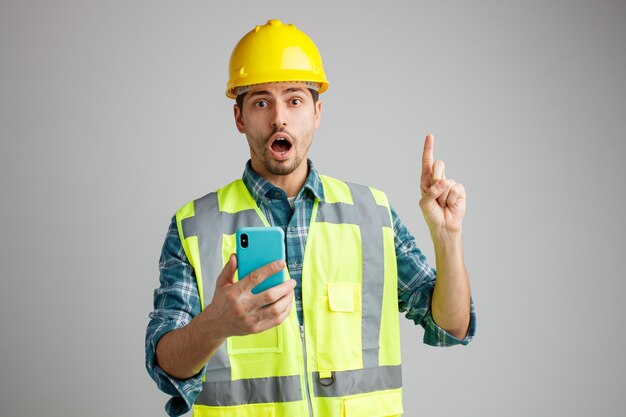 Beeindruckter junger männlicher Ingenieur mit Schutzhelm und Uniform mit Handy, der in die Kamera blickt, die nach oben zeigt, isoliert auf weißem Hintergrund