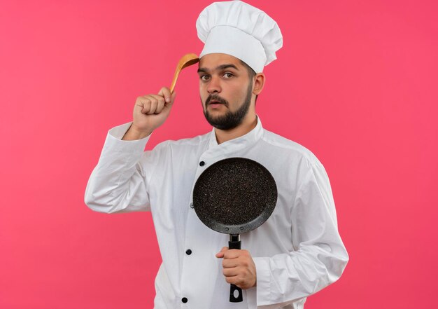 Beeindruckter junger männlicher Koch in Kochuniform, der eine Bratpfanne hält und den Kopf mit einem Löffel auf rosa Wand berührt