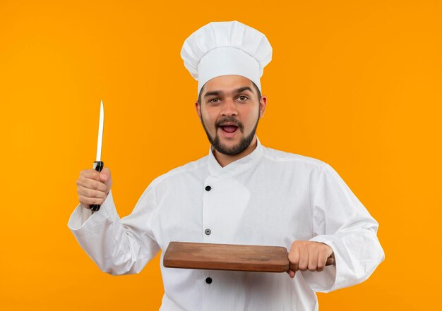 Beeindruckter junger männlicher Koch in Kochuniform mit Messer und Schneidebrett isoliert auf oranger Wand
