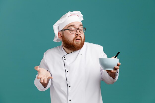 Beeindruckter junger männlicher Koch mit einheitlicher Brille und Mütze, die eine leere Schüssel mit einem Löffel darin hält und eine leere Hand zeigt, die die Schüssel isoliert auf blauem Hintergrund betrachtet
