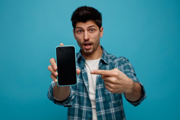 Beeindruckter junger Mann, der sein Handy in Richtung Kamera streckt und darauf zeigt, die Kamera isoliert auf blauem Hintergrund betrachtend