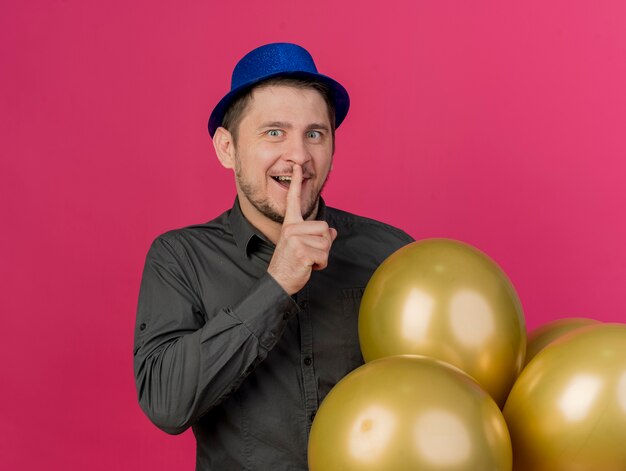 Beeindruckter junger Party-Typ, der blauen Hut trägt, der Luftballons hält, die Stille-Geste zeigen, die auf Rosa lokalisiert wird