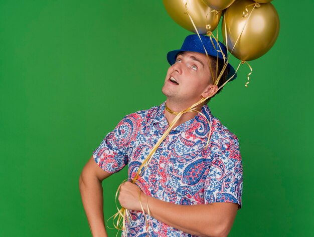 Beeindruckter junger Party-Typ, der nach oben schaut und blauen Hut trägt, der Luftballons um Hals lokalisiert auf Grün hält