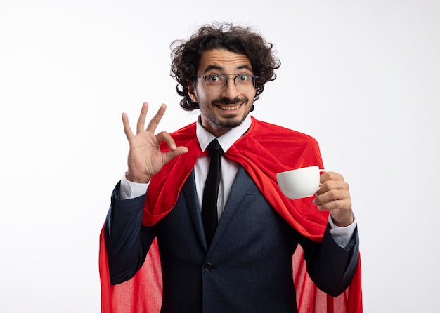 Beeindruckter junger Superheldenmann in der optischen Brille, die Anzug mit rotem Umhang trägt, hält Tasse und Gesten ok Handzeichen lokalisiert auf weißer Wand