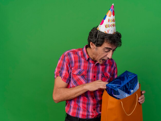 Beeindruckter kaukasischer Parteimann mittleren Alters, der Geburtstagskappe trägt, die in Papiertüte mit Geschenkverpackung lokalisiert auf grünem Hintergrund mit Kopienraum schaut