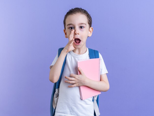 Beeindruckter kleiner Junge mit Rucksack und Notizblock, der die Hand in der Nähe des Mundes hält und auf die Kamera schaut, die isoliert auf lila Wand mit Kopierraum flüstert