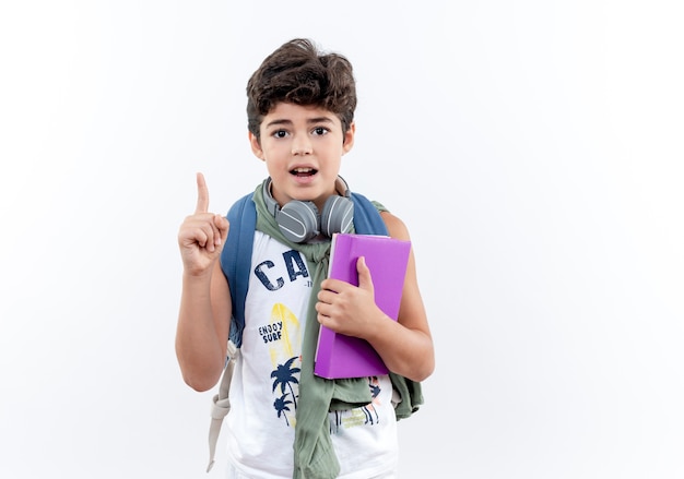 Beeindruckter kleiner Schuljunge, der Rückentasche und Kopfhörer hält Buch und Punkte oben auf weißem Hintergrund mit Kopienraum hält