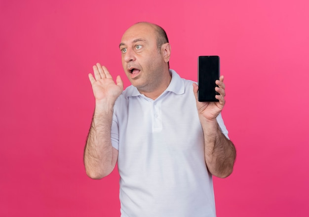 Beeindruckter lässiger reifer Geschäftsmann, der Handy hält, das Seite betrachtet und Hand in der Luft lokalisiert auf rosa Hintergrund mit Kopienraum hält