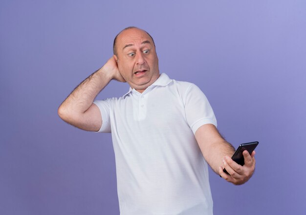 Beeindruckter lässiger reifer Geschäftsmann, der Handy hält und betrachtet und Hand hinter Kopf lokalisiert auf lila Hintergrund setzt