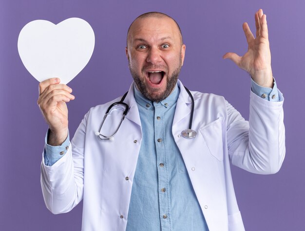 Beeindruckter männlicher Arzt mittleren Alters, der ein medizinisches Gewand und ein Stethoskop trägt, das die Hand in Herzform hält